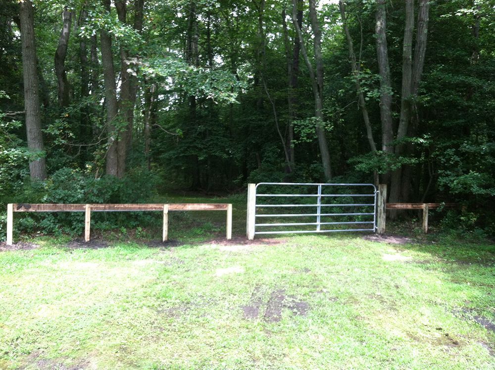 Metal gate in a clearing, leading into a wooded area.  A wooden fence extends to the left. Green grass.