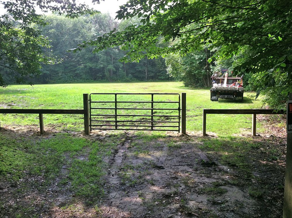 Metal gate opens to a grassy field, trees in the background, a trailer parked to the right.