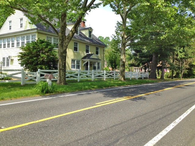Yellow house with white fence and green trees along a road.