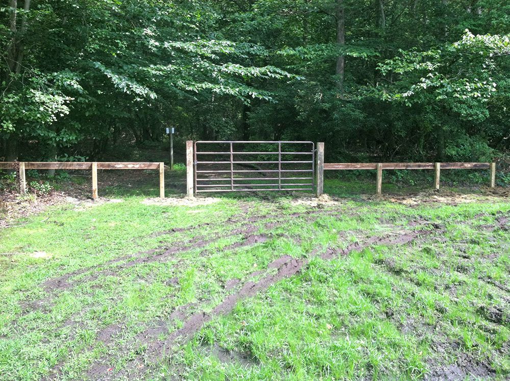 A metal gate in a wooden fence opens to a muddy field with tire tracks, forest background.