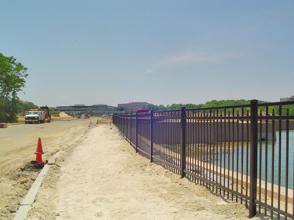 A sandy path alongside a black fence and a body of water. Construction vehicles are visible in the background on the road.