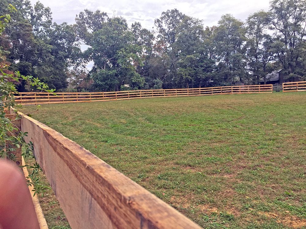 Green field enclosed by a wooden fence, with trees in the background under a cloudy sky.