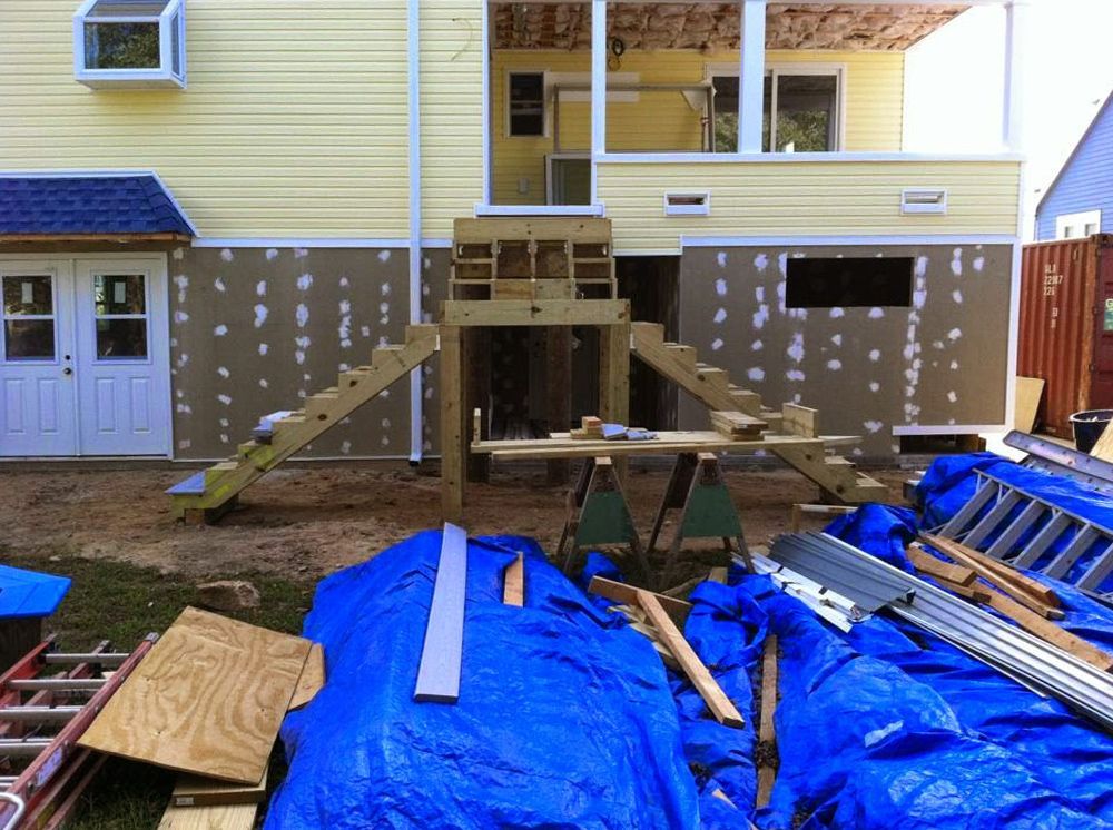 Construction site: wooden steps and framing against a yellow building with blue tarp and building materials.