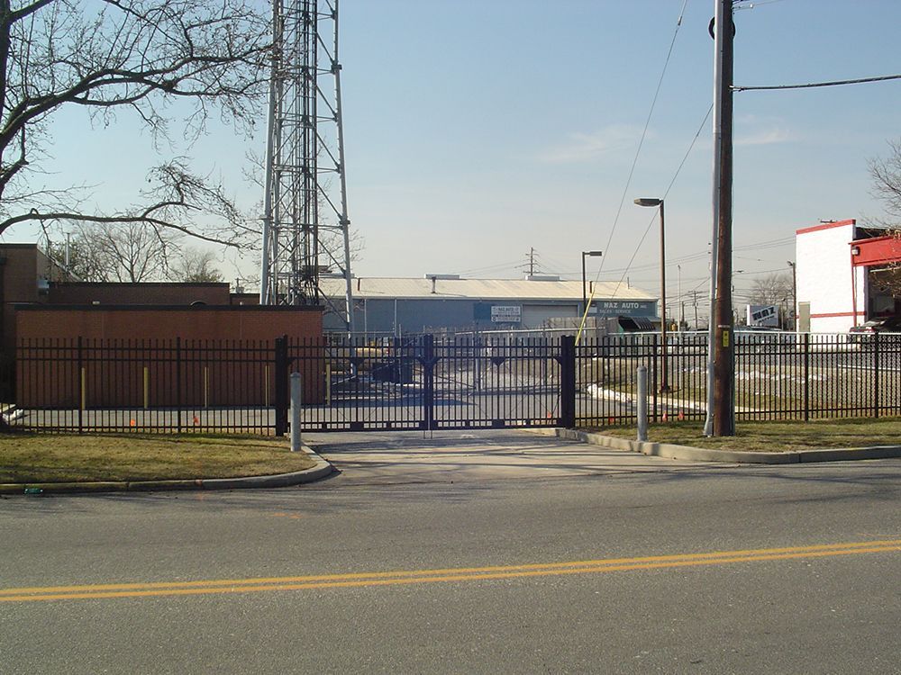 Black gate entrance to industrial complex with tall tower, light poles, and a road.