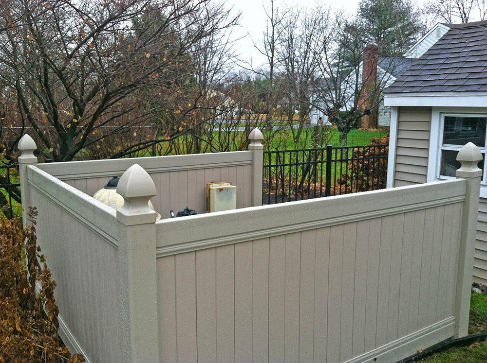 Tan vinyl fence encloses equipment in a backyard, with a black metal fence and house visible beyond.
