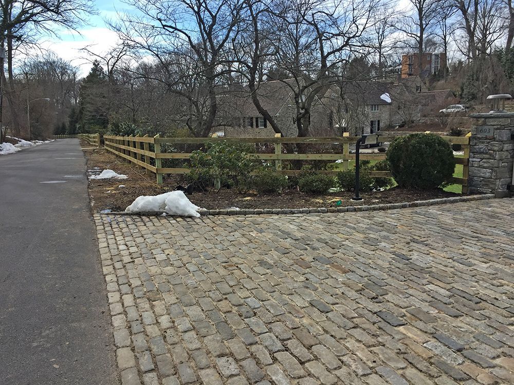 Cobblestone driveway leading to a wooden fence and house, with patches of snow and bare trees.