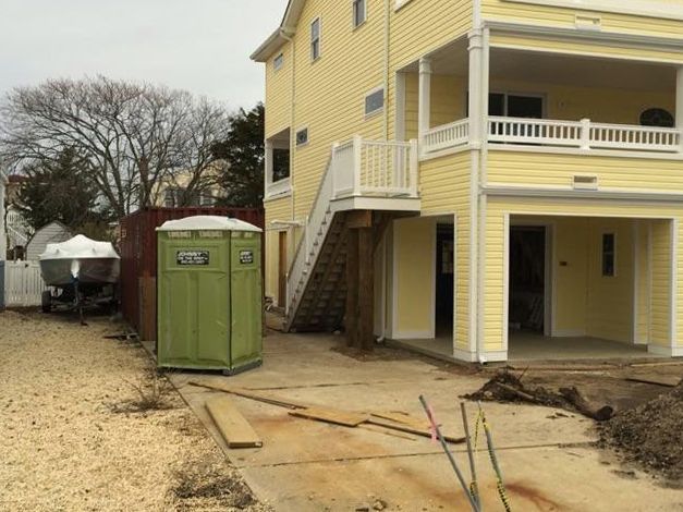 Yellow house under construction; a green porta-potty sits on a gravel driveway.