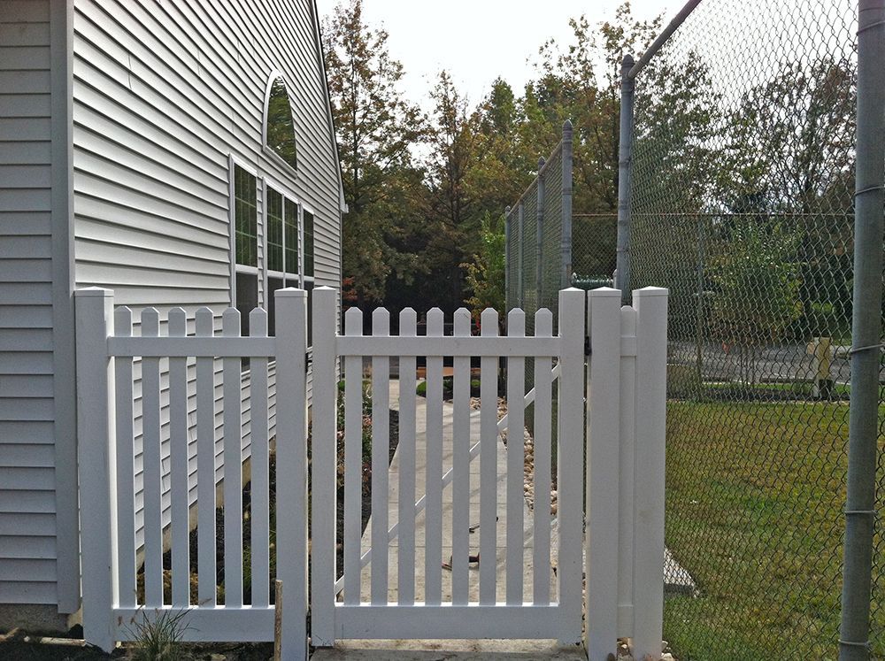 White picket fence and gate alongside a building, leading to a walkway and a chain-link fence.