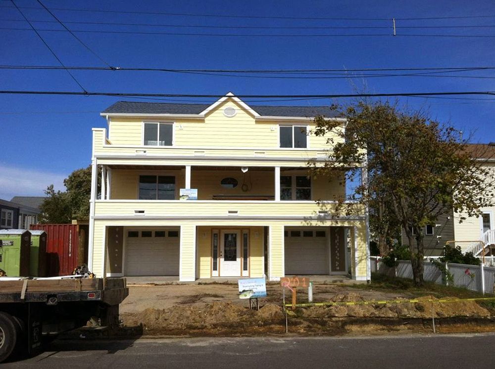 Yellow two-story house with garage doors, balcony, and front yard. Power lines and a truck in the foreground.