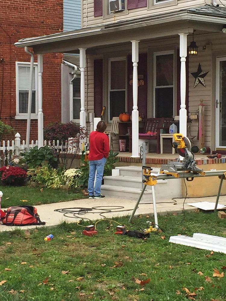Person standing on a porch with tools, preparing home improvements. Beige house with white trim, yard with tools and supplies.
