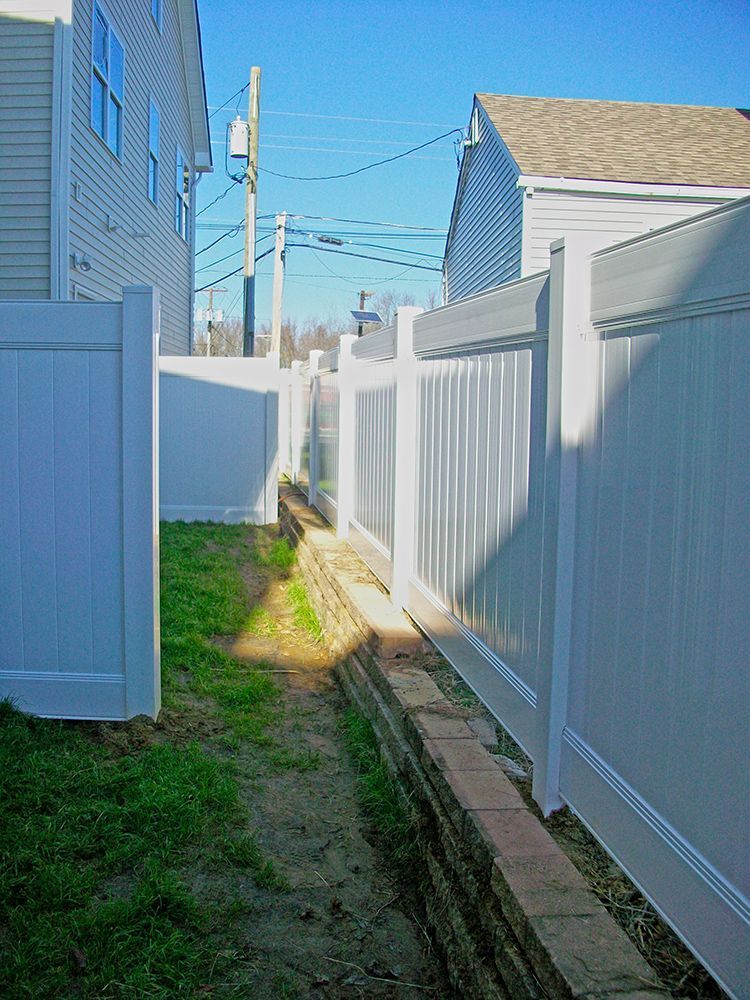 White vinyl fences border a narrow grassy pathway between two buildings on a sunny day.
