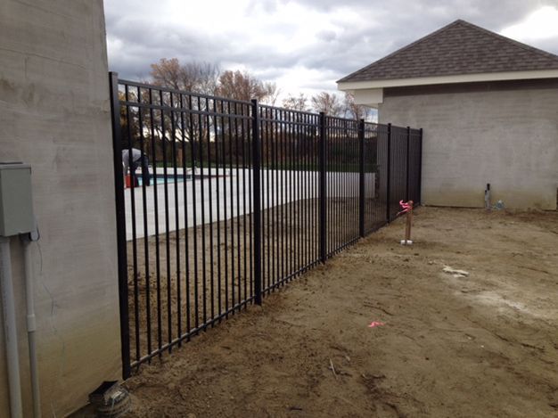 Black metal fence surrounding a pool, separating the pool from a dirt yard; a partially visible building in the background.