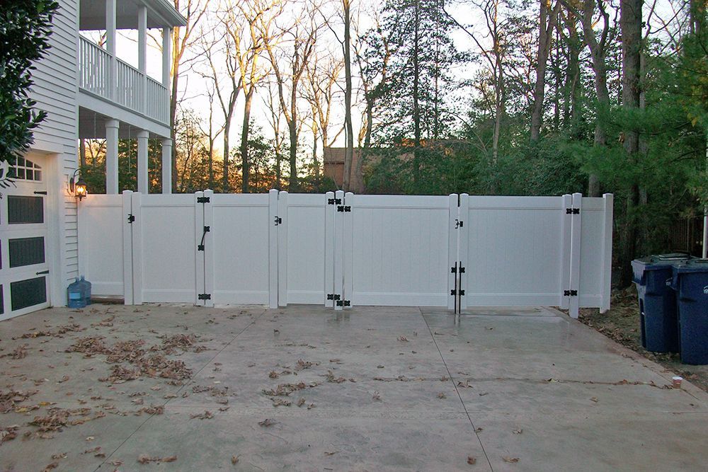 White vinyl fence with several gates in front of a house, driveway, and trees.
