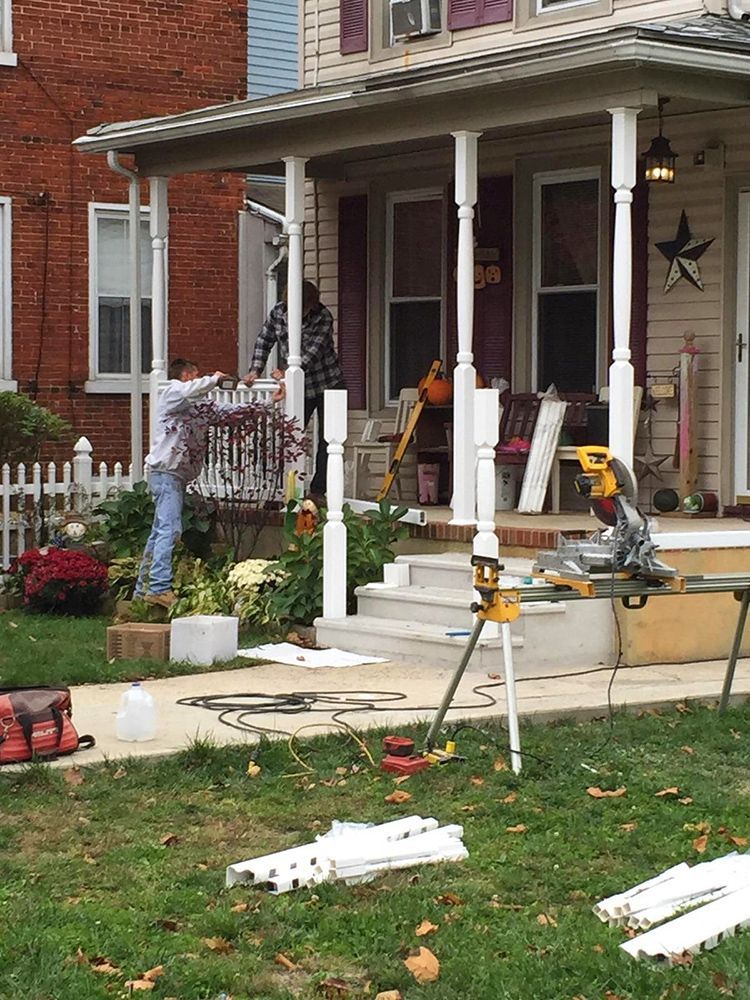Person working on a porch railing. White porch with tools, grass, and a brick house in background.