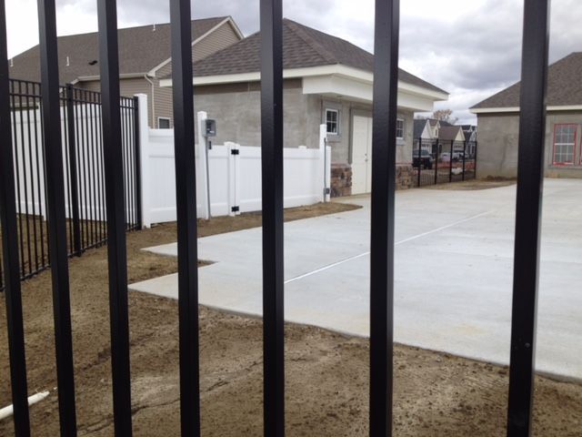Black metal fence in foreground. Behind it is a concrete patio and a white fence bordering a building with a garage.