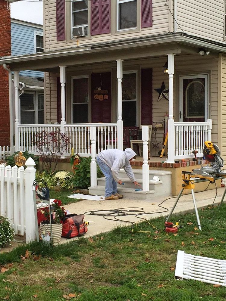Man working on a white picket fence on a porch of a two-story beige house. Tools and materials are on the ground.