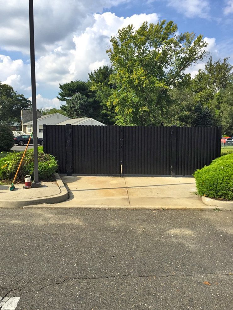 Black corrugated metal gate across a concrete driveway, flanked by bushes and trees, with a blue sky.