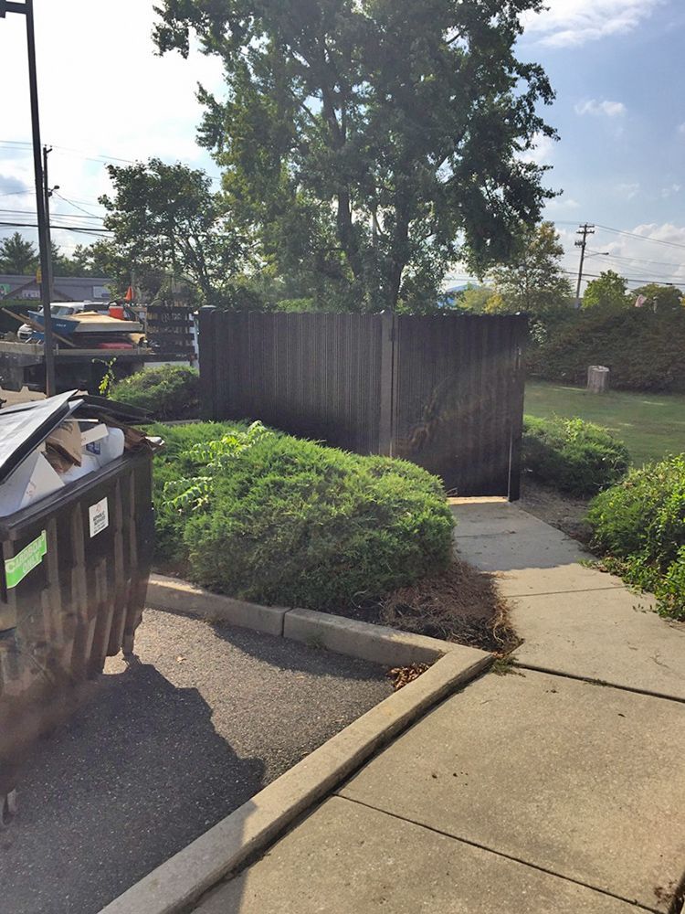 Brown dumpster and path lead to a wooden fence and shrubs. Overcast sky.