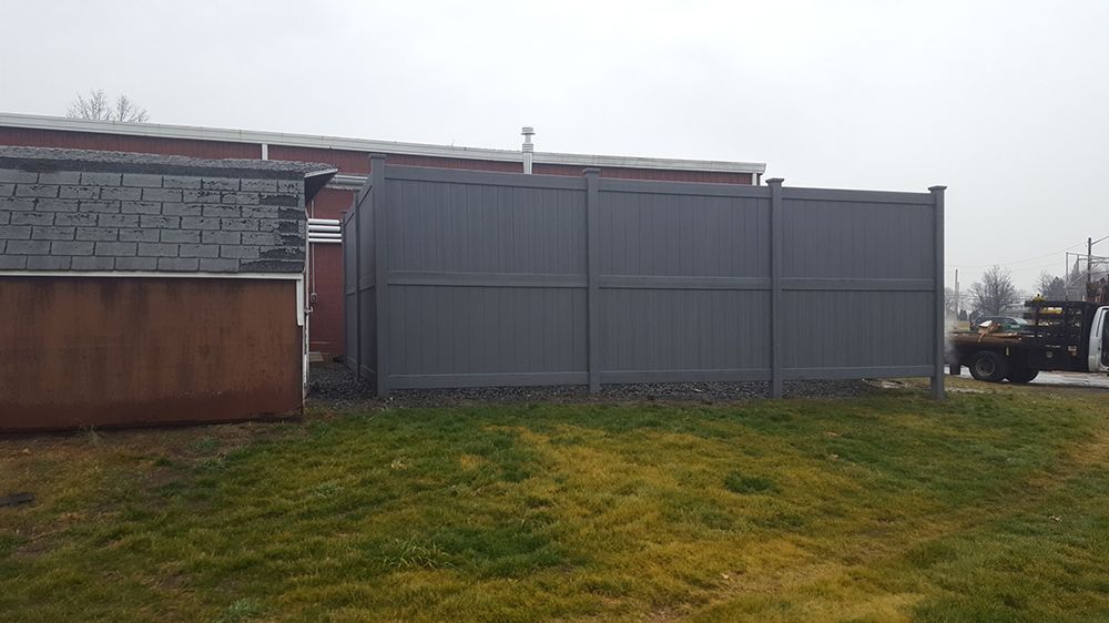 A gray privacy fence stands between a building and a grassy area, overcast sky.
