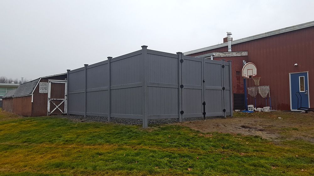 Gray fenced enclosure beside a red building and a small shed on a grassy area. Cloudy sky overhead.