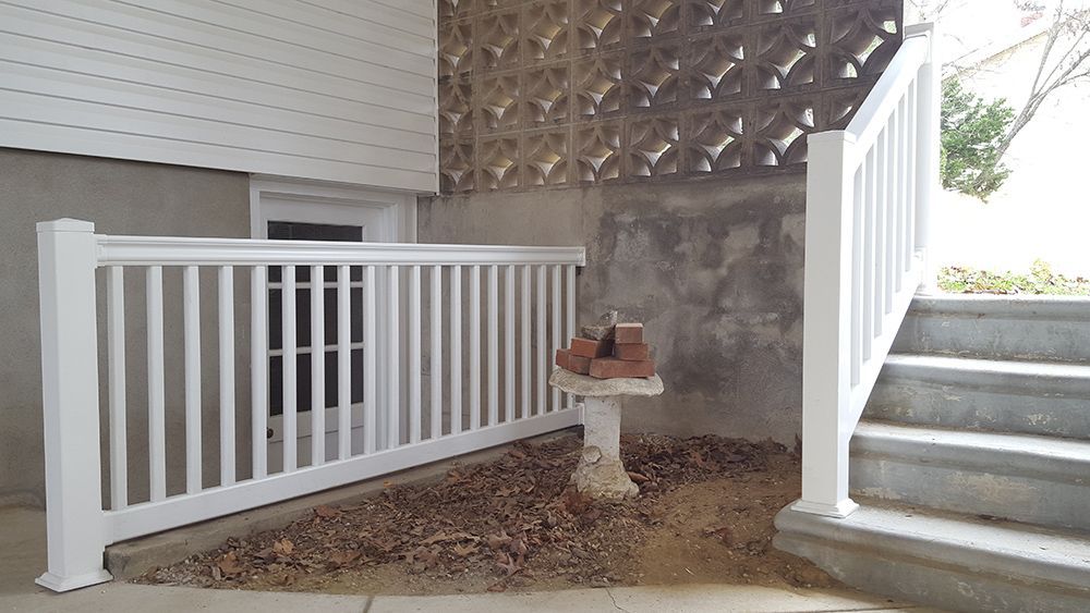 White railing surrounds a basement window and a set of concrete steps. A bird bath sits in the gravel.