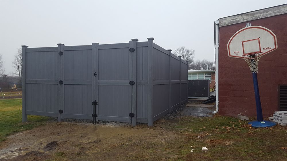 Gray fence next to a red brick building with a basketball hoop on a gloomy day.