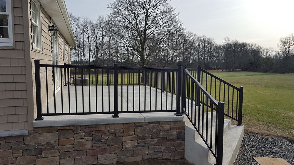 Black railing on a concrete patio next to a stone wall. A house is on the left, and a grassy field is in the background.