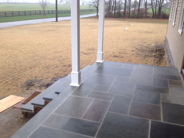 A front porch with dark gray stone tiles, white columns, and steps, overlooking a brown field.