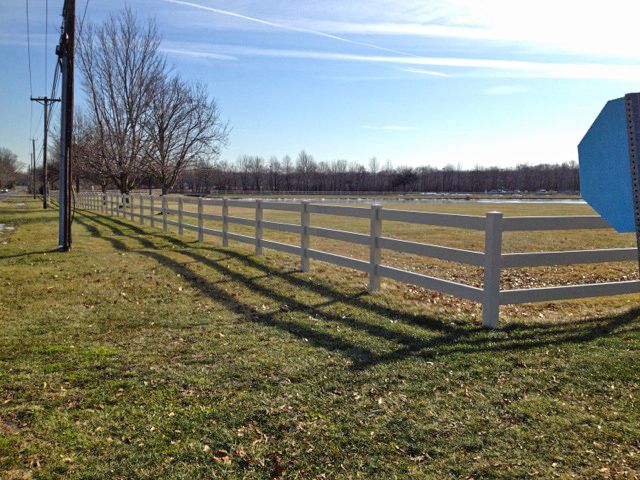 White fence running along grassy field with bare tree, power pole, and blue sky.
