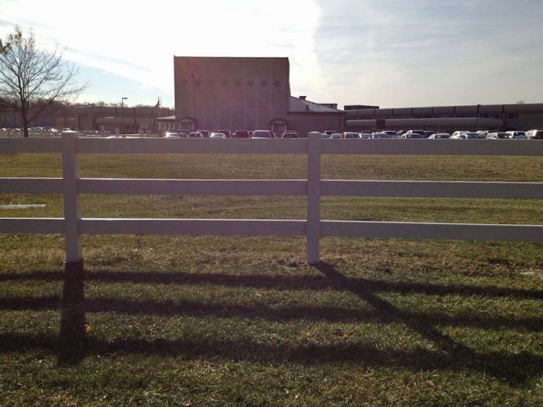 White fence in front of a grassy field, with a large industrial building and parked cars in the background.