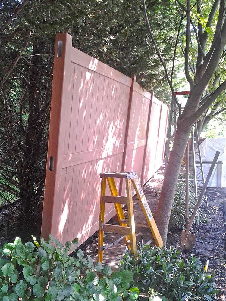 Red wooden fence being installed outdoors, with ladder and trees nearby.