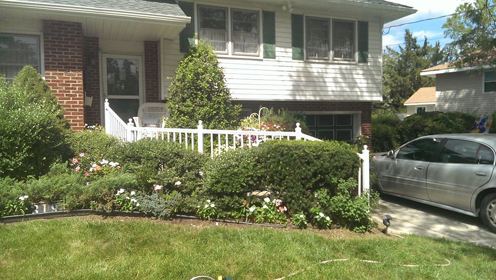 Two-story house with green shutters, white picket fence, and a car parked in the driveway. Lush garden with green foliage and flowers.