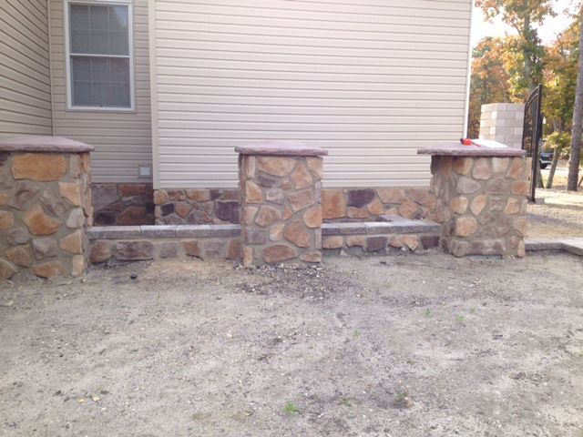 Stone pillars and wall with brown capstones, next to a beige house, against a sandy ground.