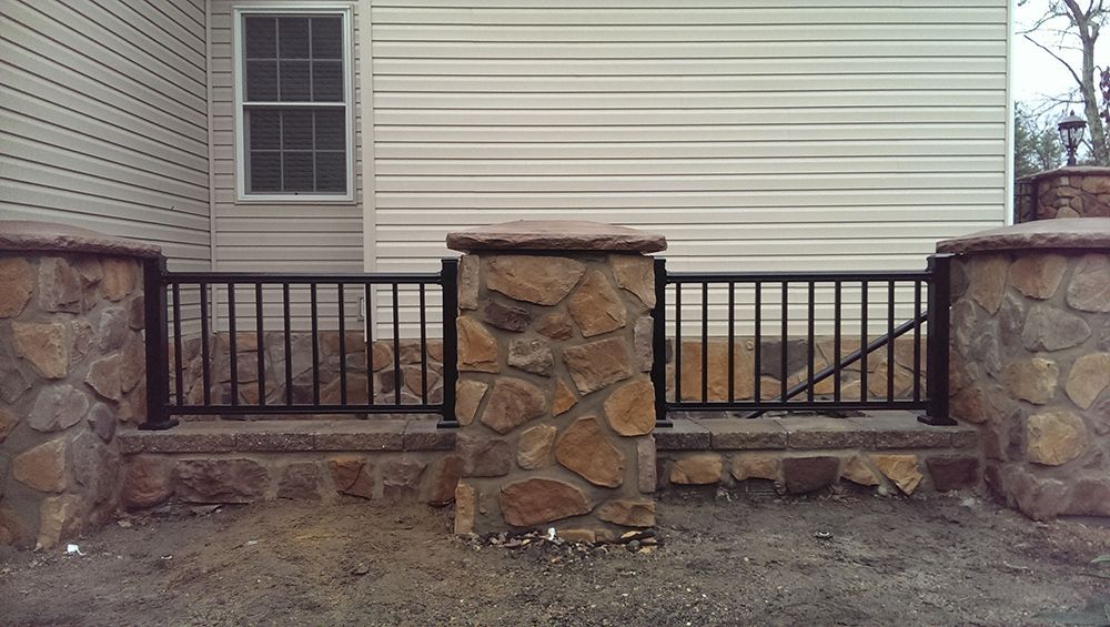 Black metal railing between stone columns in front of a house with vinyl siding and a window.