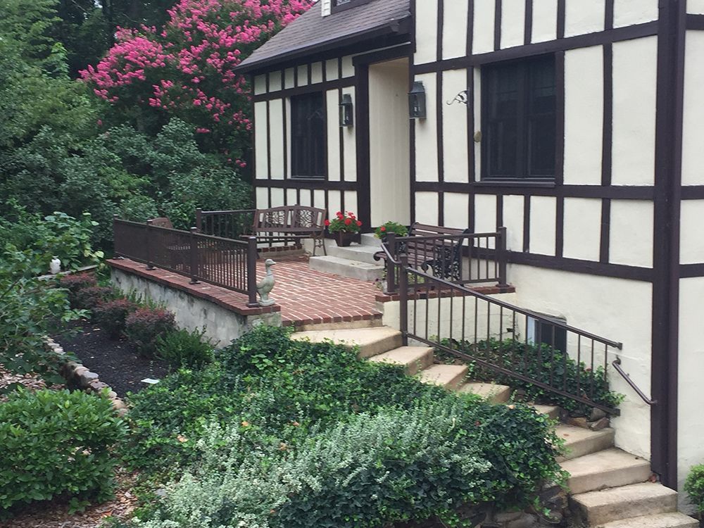 Tudor-style house entrance with steps, brick patio, brown railings, and lush green landscaping.