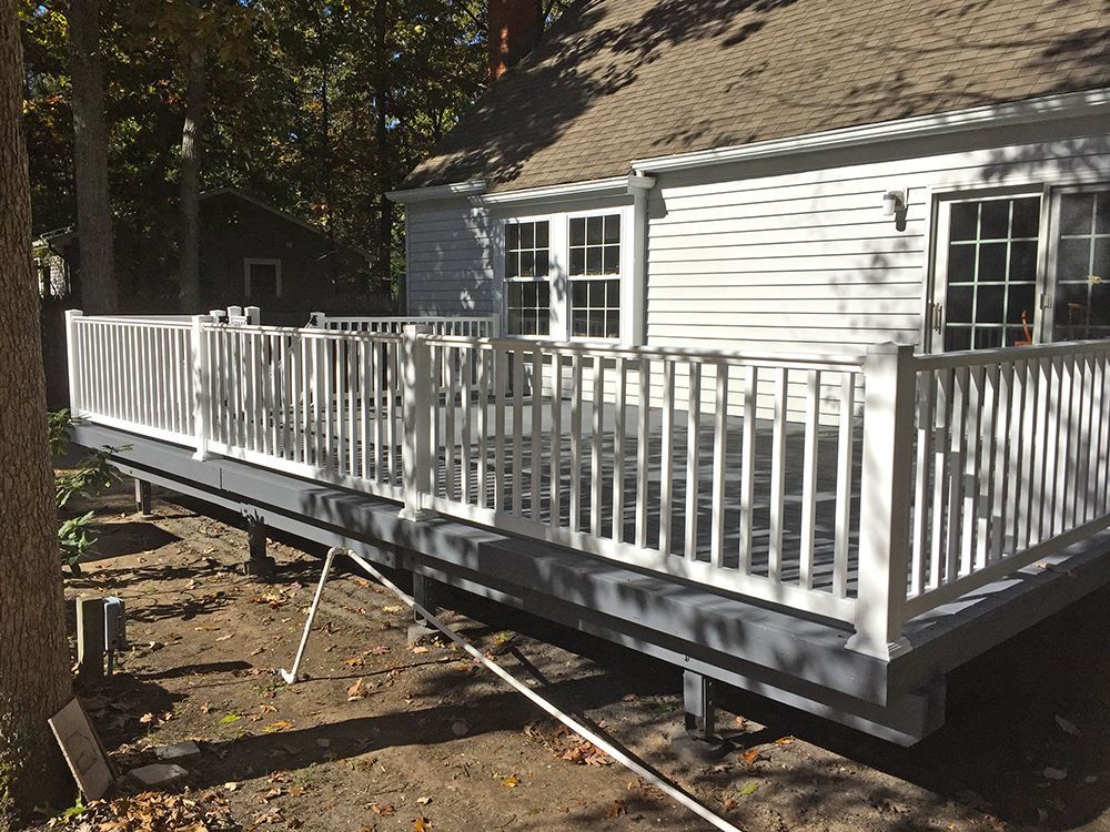 White-railed deck attached to a light-colored house. Deck is gray. Supports are visible in the dirt yard.