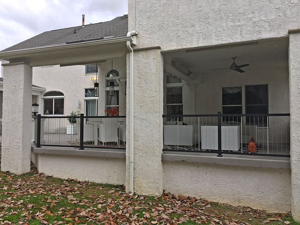 Exterior of a house with a covered patio, black railing, white stucco, and fallen leaves.
