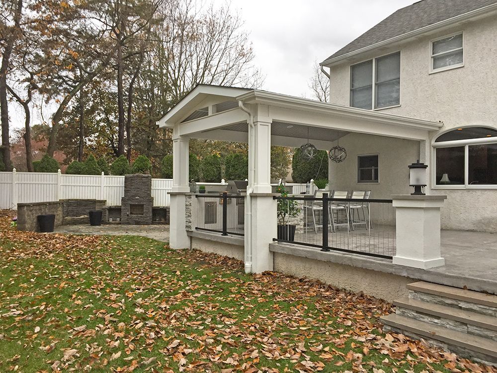 Backyard patio with a white pergola, outdoor kitchen, and a brick fireplace. Fall leaves on the grass.