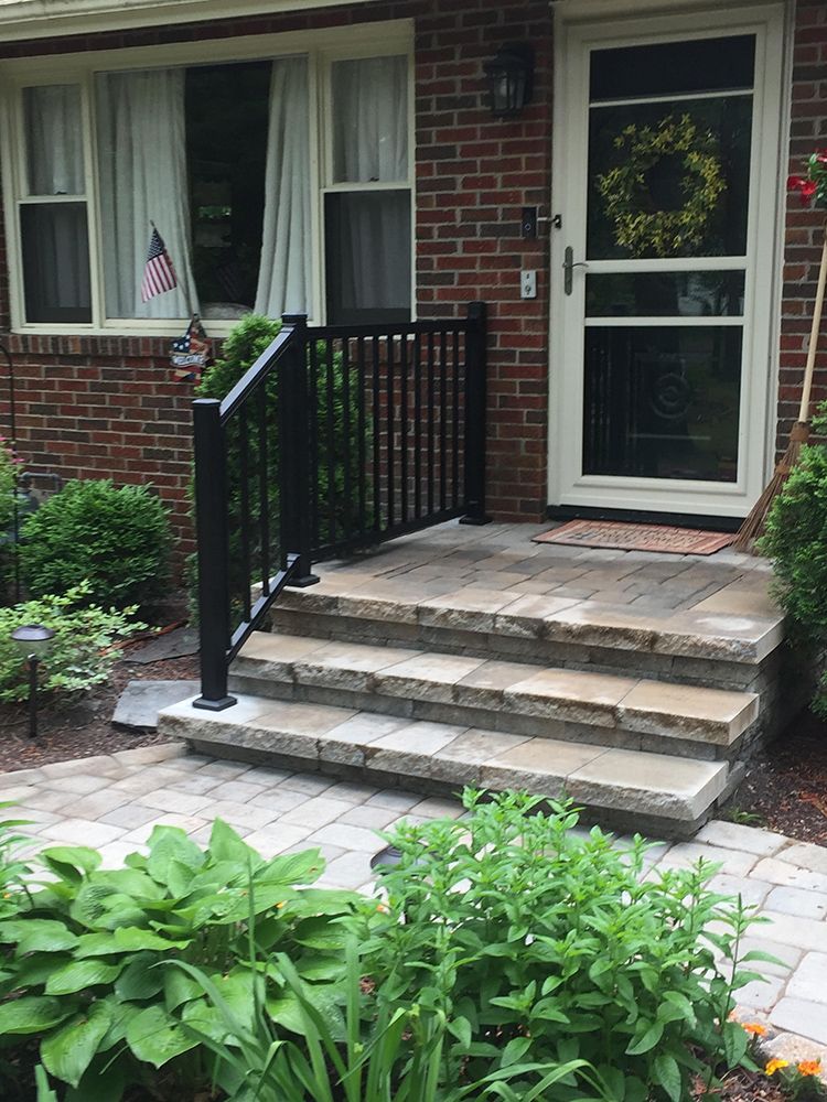 Stone steps and walkway leading to a house with black railing. Door has a screen, wreath, and is surrounded by greenery.