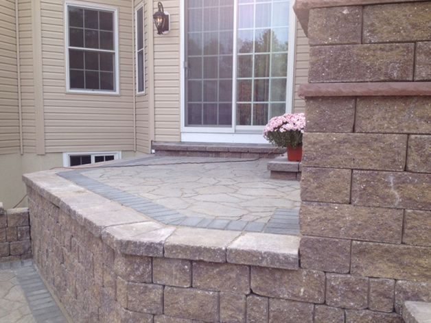 Patio with stone retaining walls and brick pavers, adjacent to a house with a sliding glass door.