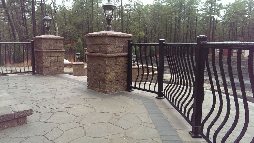 Outdoor patio with stone pavers, black metal railing, stone pillars with lamps.