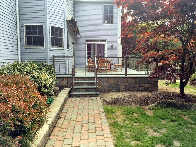 Backyard deck with stone base, wooden steps, and glass railings; red-leafed tree and brick pathway.