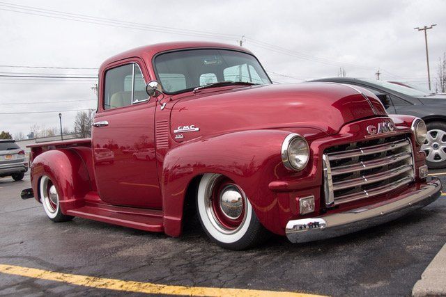Red 1950s GMC truck, low-rider style, parked on asphalt. White wall tires, chrome accents. Cloudy day.
