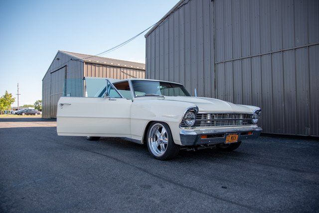 White classic Chevrolet Nova convertible with open door parked near a gray building.