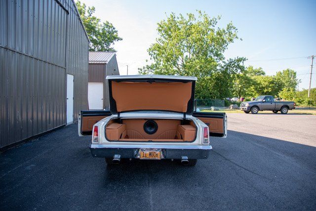 White classic car's trunk open, revealing custom brown interior and speakers, parked outdoors.