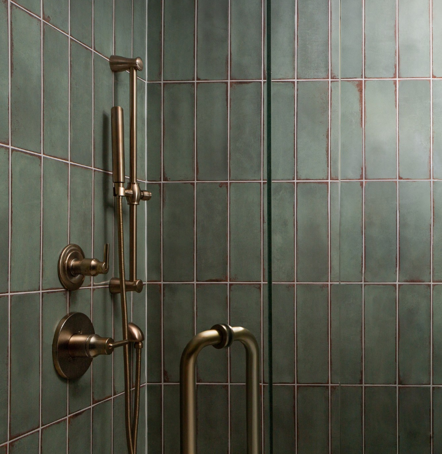 A bathroom with green tiles and a brass shower head