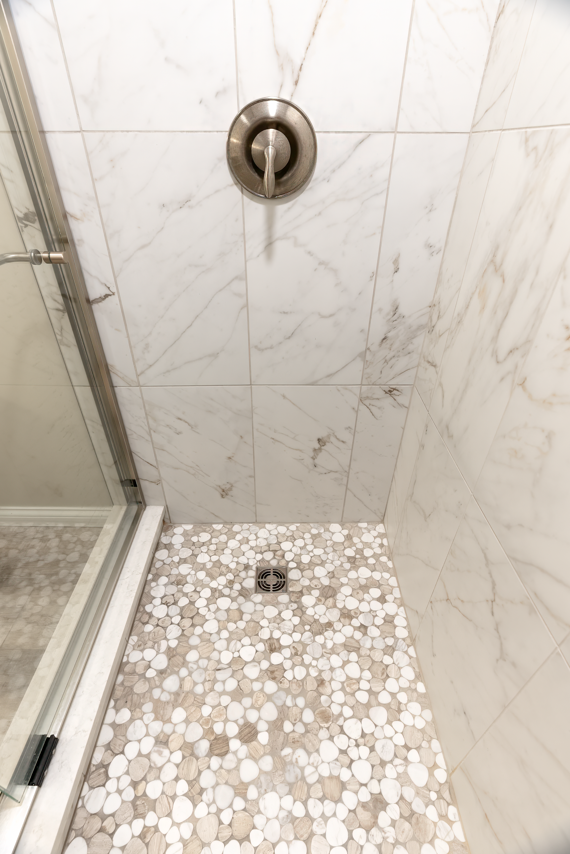 Shower with pebble tile floor and marble-like tile walls. Silver faucet and glass door.