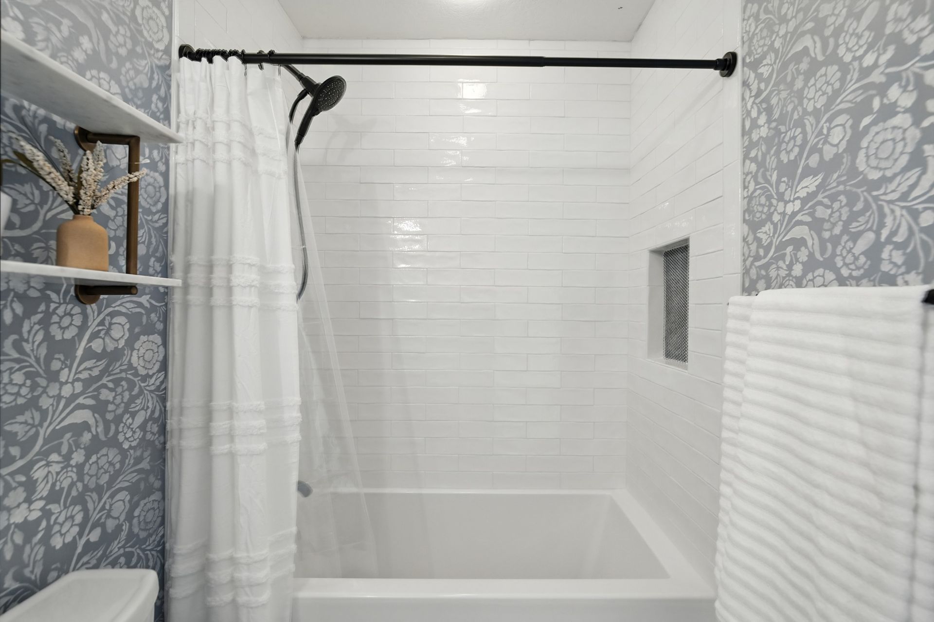 Bathroom with white tile shower, floral wallpaper, white shower curtain, and a shelf with vase.