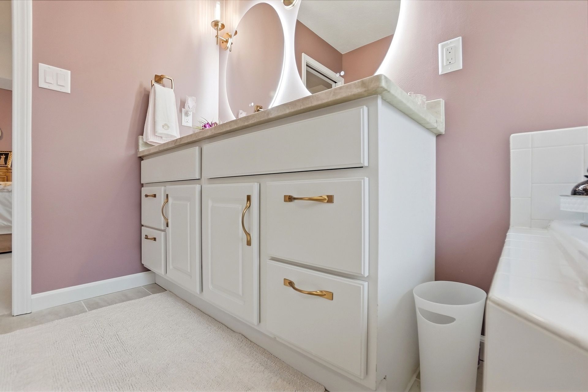White bathroom vanity with gold hardware, round mirror, and pink walls.