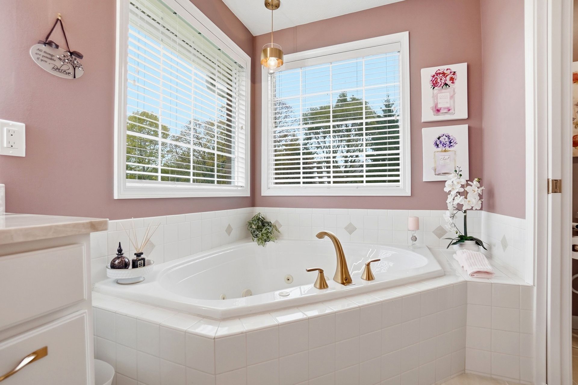 Pink-walled bathroom with white tub, gold fixtures, two windows, art, and cabinet.